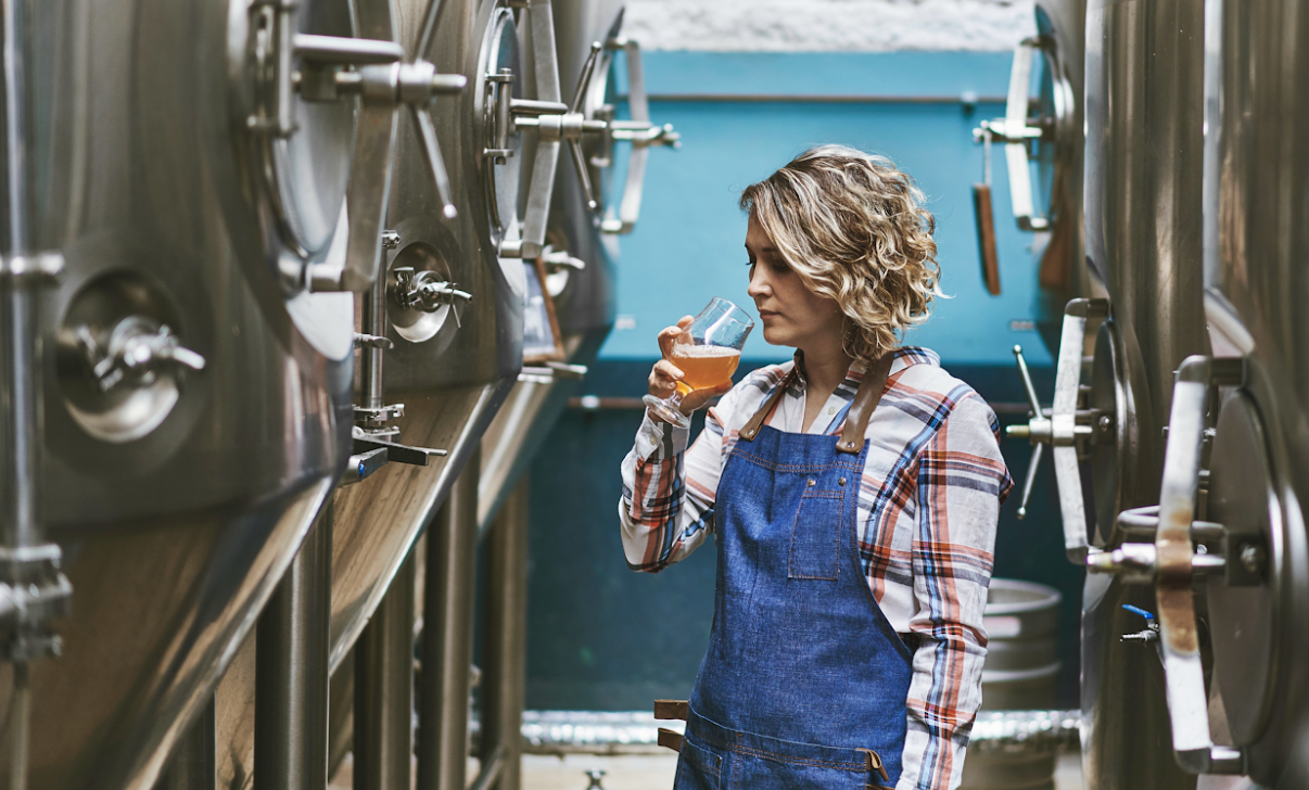 Person in a brewery wearing an apron and holding a glass of beer.