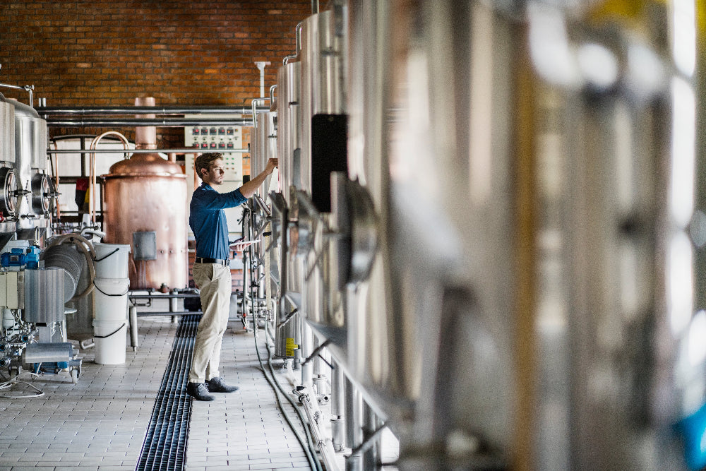 Man working in a brewery with large metal tanks and equipment.