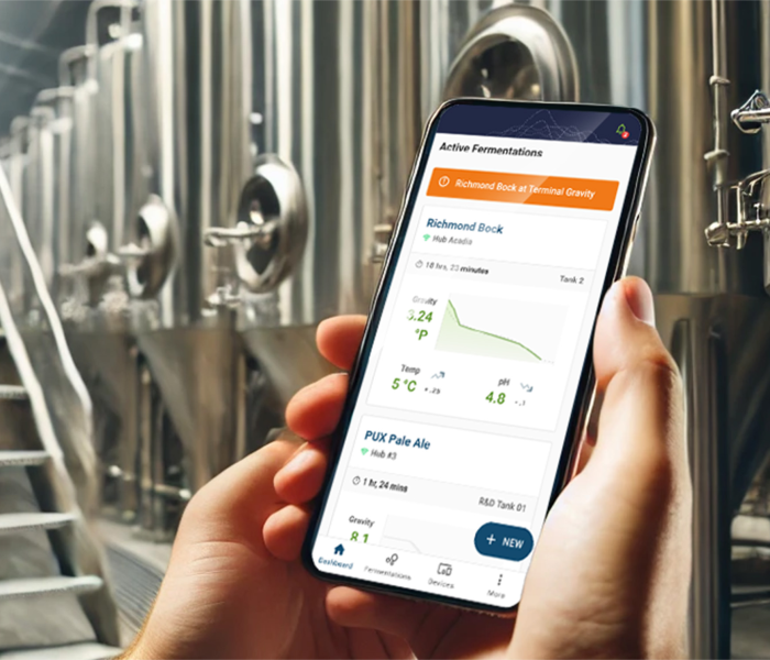 Person holding a smartphone with a brewing app in front of industrial brewing equipment