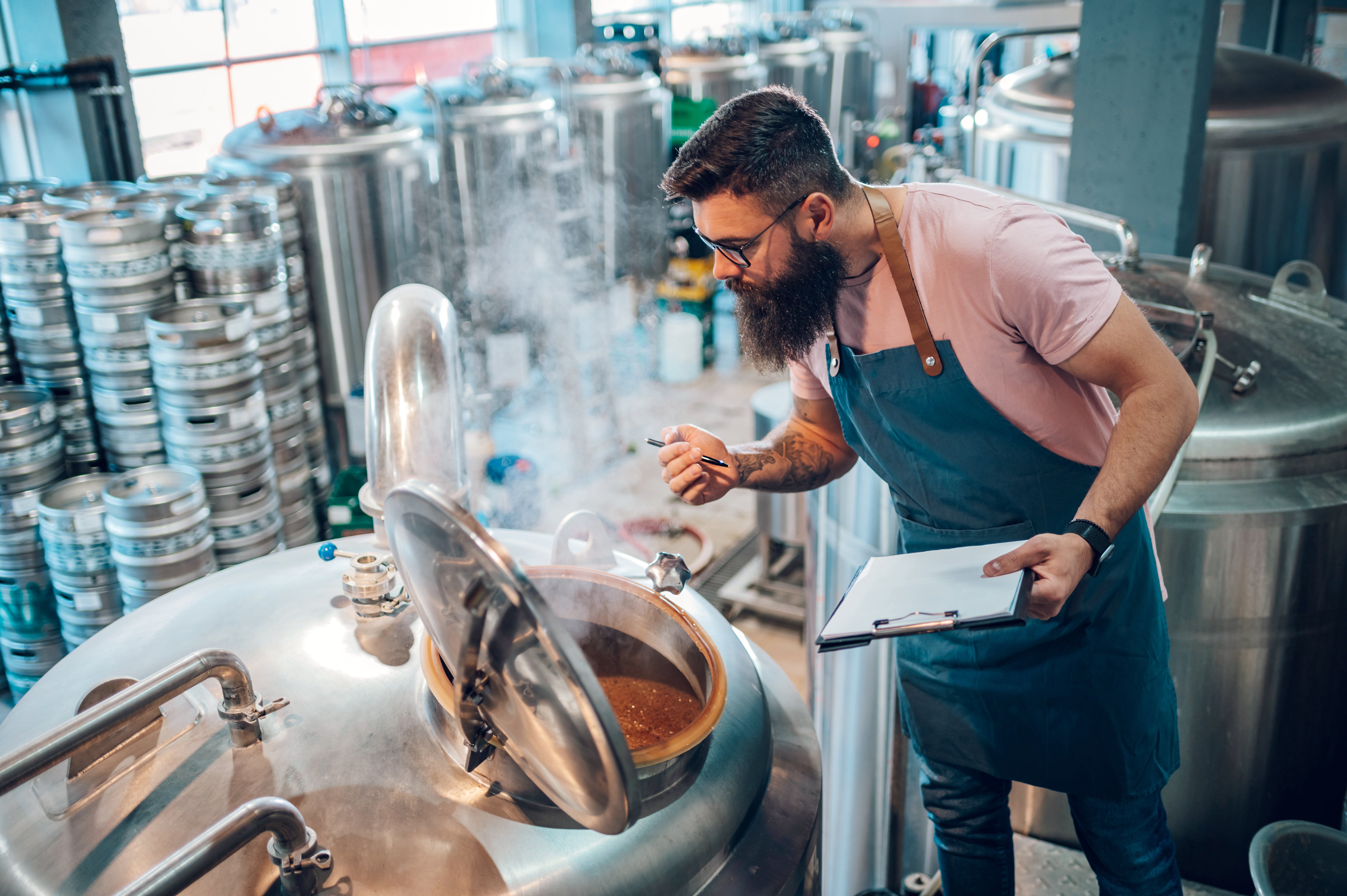 Man in a brewery checking the contents of a large metal tank with a tablet.