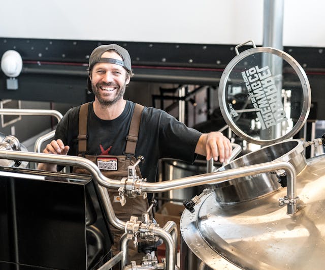 Man in a brewery setting with large metal tanks and a sign in the background